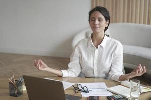 Mujer italiana tranquila con los ojos cerrados meditando en el lugar de trabajo mientras se sienta en la mesa con una laptop foto