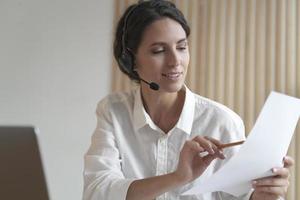 Positive Spanish businesswoman in headset sits at desk having video conference online with partners photo