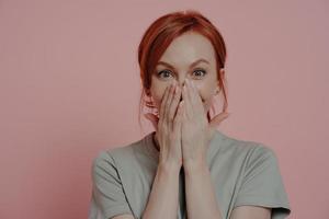 Young pretty ginger woman covering mouth with both hands, being in shock and disbelief photo