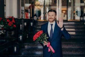 Attractive man waiting on street in elegant dark blue suit with red rose flower bouquet photo