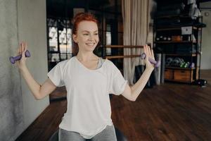 Happy redhead young woman sitting on silver fitball and exercising with small dumbbells
