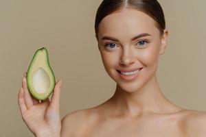 Close up shot of topless young Caucasian female model has dark combed hair smiles gently holds half of avocado stands shirtless indoor cares about skin and complexion. Beauty wellness concept photo
