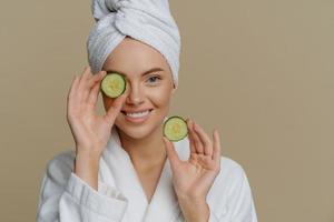 Skin care natural beauty and cosmetology concept. Positive refreshed beautiful woman holds slices of fresh cucumber makes mask for face skin dressed in domestic robe wrapped bath towel on head
