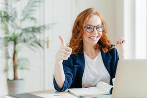 Positive redhead woman keeps thumb raised, demonstrates like gesture, satisfied with good work of colleague, updates software on modern gadget, searches data on website, poses in coworking space photo