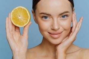 Close up shot of nice looking young woman touches face gently has well cared complexion healthy glowing skin stands bare shoulders holds half of juicy lemon isolated over blue studio background. photo
