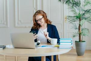 mujer pelirroja sonriente enfocada en un celular moderno, feliz de recibir mensajes de texto, posa en el lugar de trabajo, bebe café, trabaja en un proyecto financiero y se prepara para una videoconferencia con un socio comercial foto