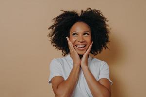 Joyful young dark skinned woman feeling happy, smiling at camera and touching cheeks with both hands photo