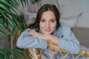 Attractive young woman with dark hair, nice make up looks directly at camera, leans on back of wicker furniture, domestic atmosphere, green plant, has confident expression. People and rest concept photo