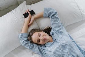 Shot of pretty brunette student learns language via mobile app, listens audio with earphones, has free time, dressed in casual outfit, poses on comfortable white bed, has thoughtful look aside photo