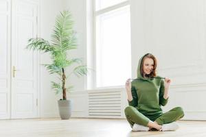 Positive female wears hoody, sits in lotus pose, has fun after doing sport, glad to have morning yoga training, fitness workout, models on floor, feels happy. People, energy, exercising concept