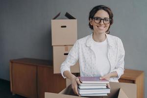 Happy nerd in glasses is stacking books into boxes. Young spanish woman packing carton container. photo