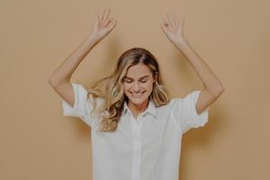 Carefree lovely female in white shirt with blonde hair enjoying nice day, keeping eyes closed and raising arms