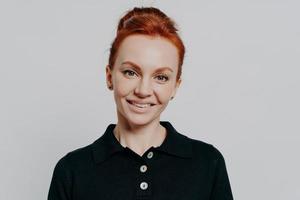 Smiling young woman with red hair in bun looking at camera isolated over grey background photo