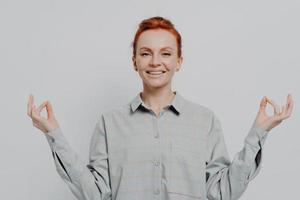 Peaceful young smiling redhead woman holding hands in mudra isolated on grey studio background