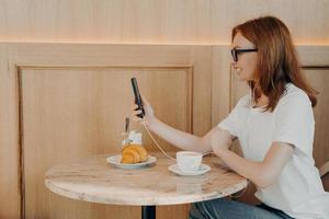 una joven pelirroja feliz usa una conexión de roaming sentada a la mesa en una cafetería