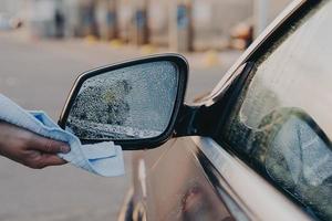 Male hand cleaning car outdoors with rag, wiping water with microfiber cloth on side rearview mirror photo