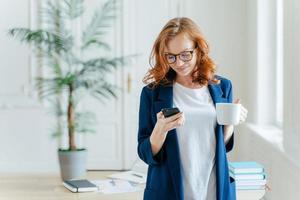 Successful female CEO reads notification on modern cell phone, has coffee break, dressed in formal clothes, starts work day from hot drink, poses over office interior, checks email box online photo