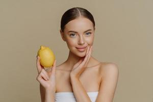 Portrait of beautiful brunette European woman holds fresh yellow lemon for making natural cream mask poses wrapped in towel isolated over brown background. Skin care hygiene and beauty concept
