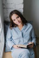 Relaxed young of beautiful in casual clothes, sits at window near brick wall, back to soft pillow, reads interesting detective story, looks directly at camera, wears casual costume. People and reading photo