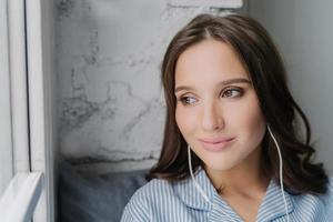 Headshot of attractive female meloman enjoys music in earphones with high volume, likes peaceful atmosphere, looks thoughtfully at window, listens favourite song. People, rest, technology concept photo