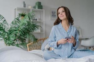 Shot of pretty brunette student learns language via mobile app, listens audio with earphones, has free time, dressed in casual outfit, poses on comfortable white bed, has thoughtful look aside photo