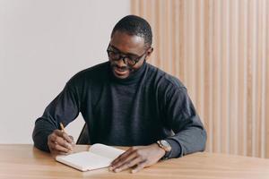 Young african american man entrepreneur in glasses taking notes in agenda photo