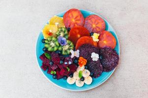 Top view of tropical fruit assortment on plate against white background. Fresh slices of dragon fruit, pitaya, kiwi, banana, blueberry decorated with small pansy. Bright colored healthy fruit