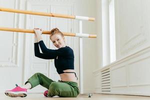 Shot of beautiful ginger woman does pilates exercises on handrail, sits on floor, stretches herself, poses in dancing studio, being in good body shape, dressed in sportswear. Sport and health care photo