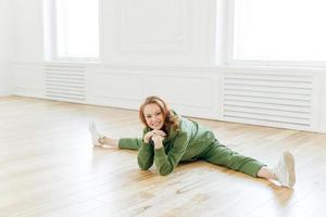 Glad refreshed sporty red haired woman has work out in empty hall, demonstrates flexibility exercise, keeps hands under chin, dressed in green tracksuit and sneakers. Yoga and fitness concept.