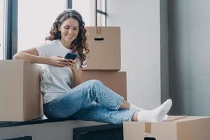 Happy spanish woman is sitting on windowsill texting on phone and unpacking boxes. photo