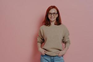 Happy redhaired female student in spectacles smiling at camera, isolated on pink background photo
