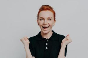 Happy young ginger girl being excited by great news, raising hands with clenched fists