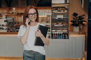 Woman entrepreneur with ginger hair has telephone conversation holds laptop photo
