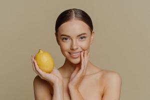 Portrait of charming half naked woman with dark combed hair holds fresh whole lemon uses fruit for natural cosmetics poses against brown background with naked body. People beauty wellness concept photo