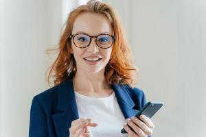Headshot of female banker with pleasant appearance and red hair, does banking online on cell phone, checks notification on financial website, wears transparent glasses and elegant formal suit photo