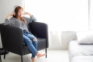 Beautiful asian young woman enjoy and fun listen music with headphone sitting on chair in the bedroom, girl relax with earphone, leisure and technology concept. photo