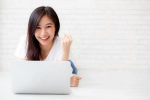 hermoso retrato de una joven asiática emocionada y contenta del éxito con una computadora portátil, una chica acostada trabajando en un fondo de ladrillos de cemento, un concepto de negocio independiente de carrera. foto