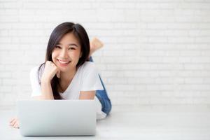 Beautiful asian woman working online laptop lying on floor brick cement background, freelance girl using notebook computer with connect to internet for distance job, business and lifestyle concept. photo