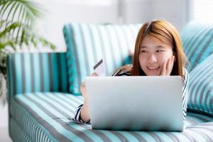 hermoso retrato de una joven asiática que miente a los usuarios de la tarjeta de crédito con una computadora portátil, una chica contenta que compra en línea y paga con una computadora portátil en el sofá, concepto de estilo de vida. foto