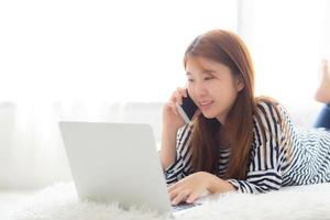 Beautiful asian young woman lying on bed using laptop and talking mobile smart phone at bedroom for leisure and relax, freelance with girl working notebook, communication concept. photo