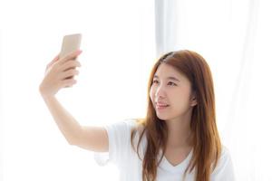 Beautiful portrait young asian woman taking a selfie with smart mobile phone on bedroom, girl is photographing with happy and smile with fun, lifestyle concept. photo