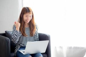 hermosa de retrato mujer joven asiática emocionada y contenta de éxito con una computadora portátil en la silla en el dormitorio, chica trabajando, concepto de negocio independiente de carrera.