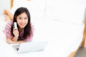 hermoso retrato joven mujer asiática que miente a los usuarios tarjeta de crédito con computadora portátil, chica contenta comprando en línea y pago con computadora portátil en la cama, concepto de estilo de vida. foto