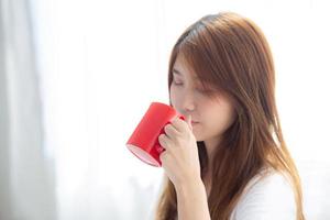 hermoso retrato de una joven asiática con una taza de café de pie en el fondo de la ventana de la cortina en el dormitorio, la chica se relaja por la mañana en casa, el concepto de estilo de vida. foto