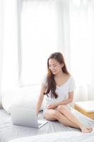 retrato de una hermosa joven asiática sentada en la cama usando una computadora portátil en el dormitorio para el ocio y la relajación, independiente con un cuaderno de trabajo para niñas, comunicación y concepto de estilo de vida.