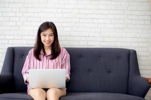 Beautiful of portrait asian young woman working online laptop sitting on sofa at living room, girl using notebook computer with connect to internet for distance job, business and success concept. photo