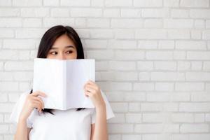 beautiful portrait young asian woman happy hiding behind open the book with cement or concrete background, girl standing reading for learning, education and knowledge concept. photo