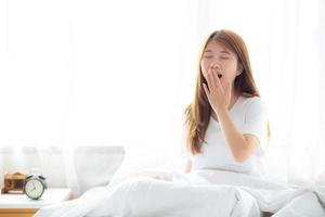 Beautiful of portrait young asian woman yawn sitting with sleep on bed at bedroom, girl wake up after resting and leisure with wellness, lifestyle concept. photo