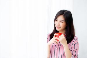 hermoso retrato de una joven asiática con una taza de café de pie en el fondo de la ventana de la cortina en el dormitorio, la chica se relaja por la mañana en casa, el concepto de estilo de vida. foto