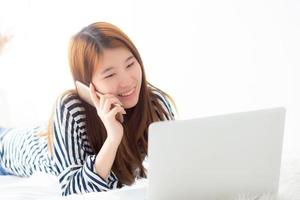 hermosa mujer joven asiática acostada en la cama usando una computadora portátil y hablando de un teléfono inteligente móvil en el dormitorio para el ocio y la relajación, independiente con un cuaderno de trabajo para niñas, concepto de comunicación. foto